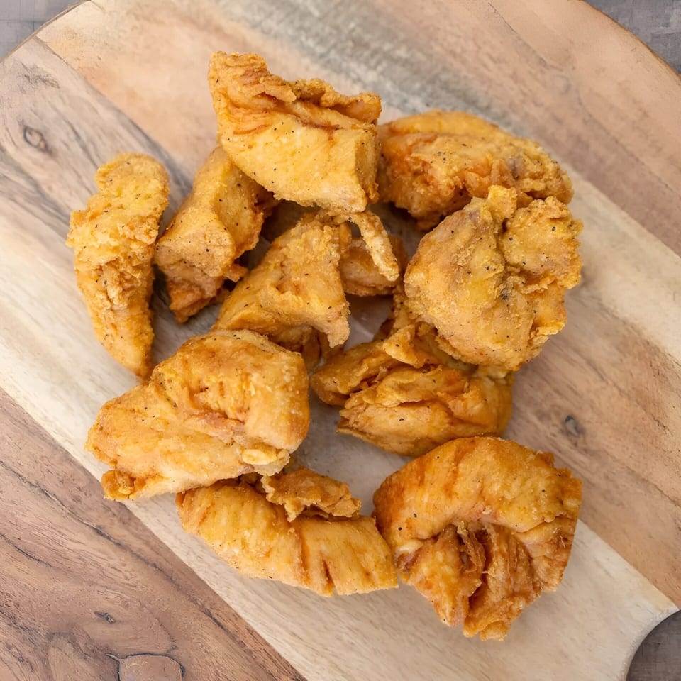 Golden-brown fried chicken pieces, seasoned with pepper, piled on a rustic wooden cutting board.