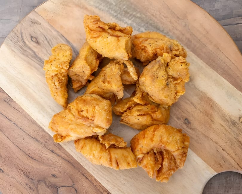 Golden-brown fried chicken pieces, seasoned with pepper, piled on a rustic wooden cutting board.