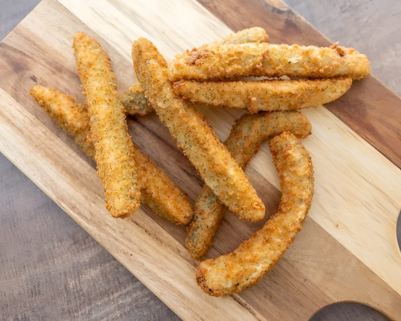 Golden brown fried pickle spears, coated in breading and dill, arranged on a wooden cutting board.