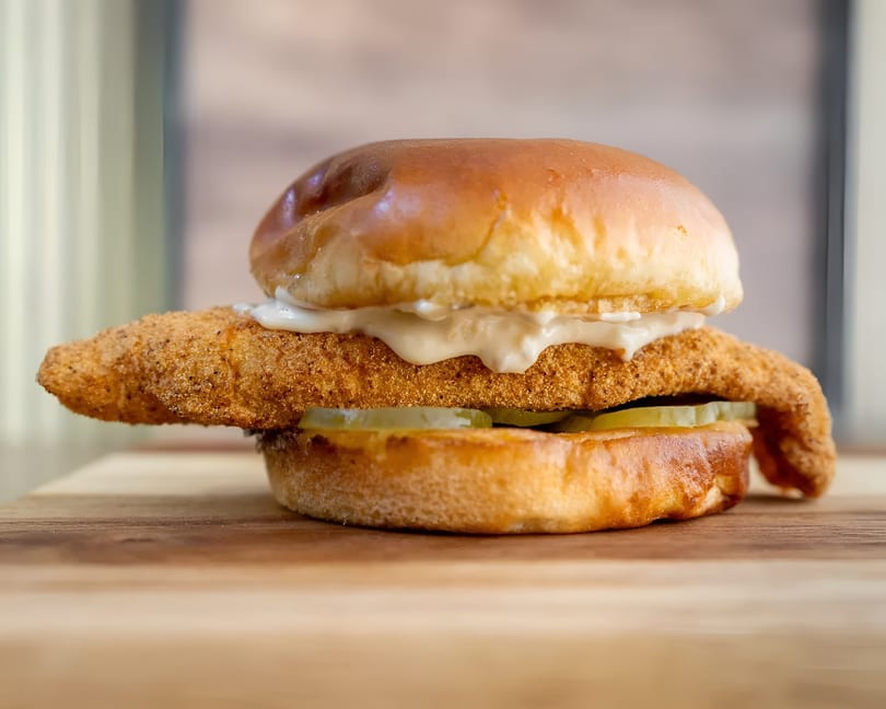 Crispy fried fish sandwich with tartar sauce and pickles on a brioche bun, on a wooden board.
