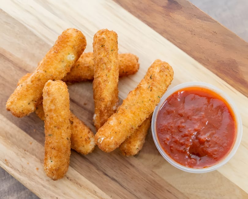 Golden-brown mozzarella sticks with herbs and a side of marinara sauce on a wooden board.