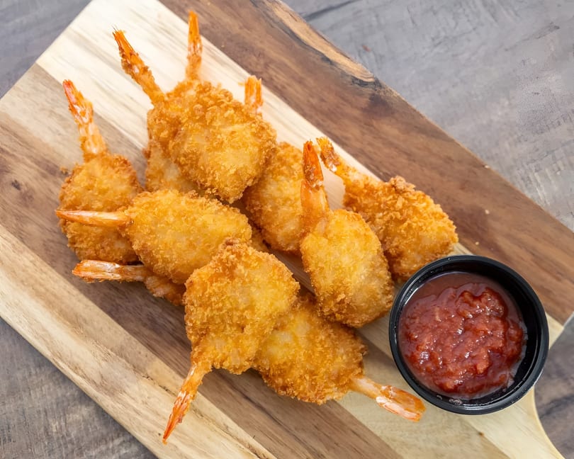 Crispy fried shrimp with tails on, served with a side of red dipping sauce on a wooden board.