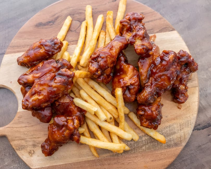 Sweet and sticky BBQ chicken wings with a side of golden french fries on a wooden board.