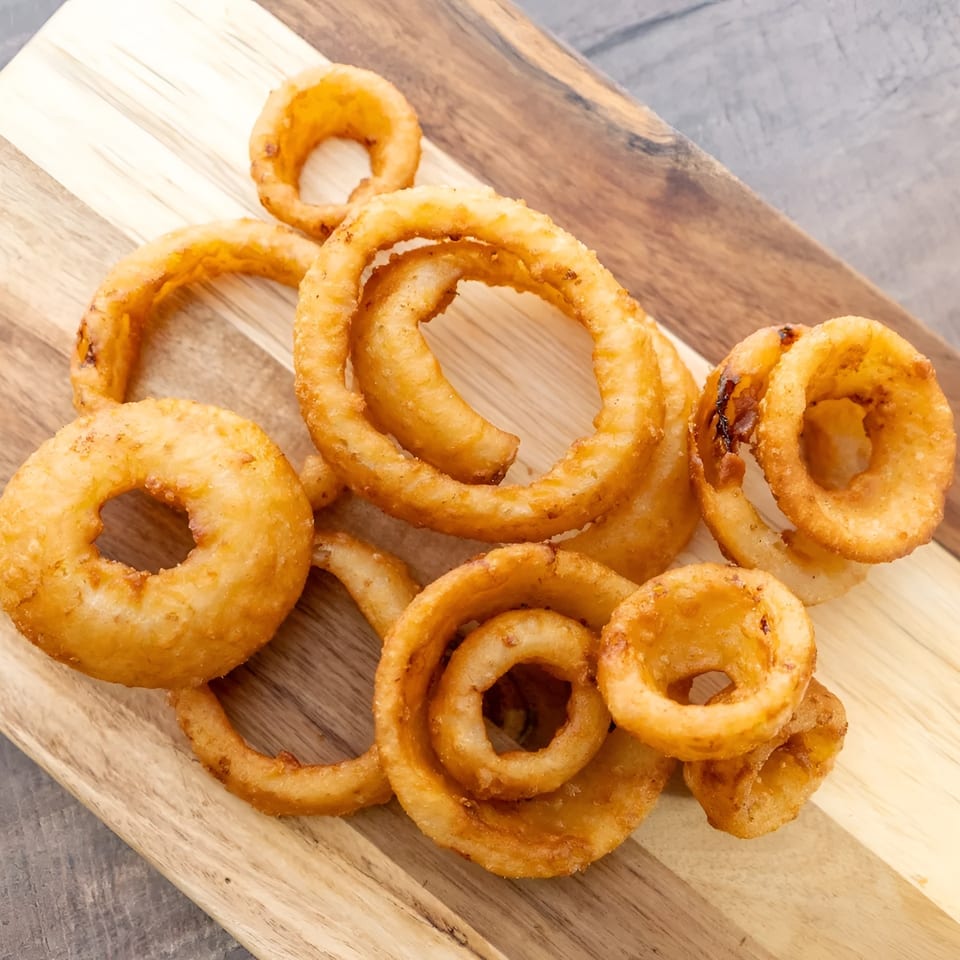 Golden brown onion rings, some curly and some round, piled on a wooden cutting board.