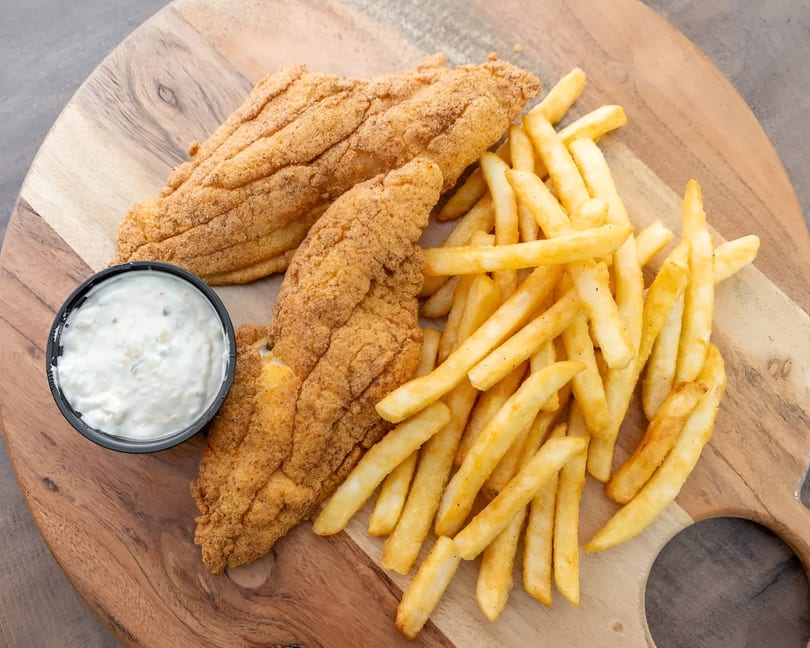 Two pieces of fried fish, a side of french fries, and a cup of tartar sauce on a wooden board.