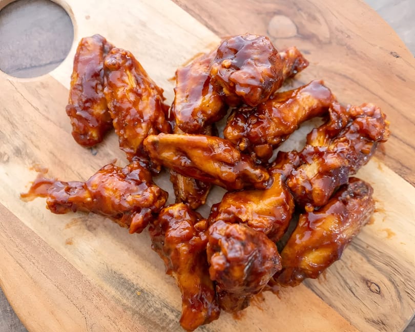Pile of sticky, glistening barbecue chicken wings on a light wooden cutting board.