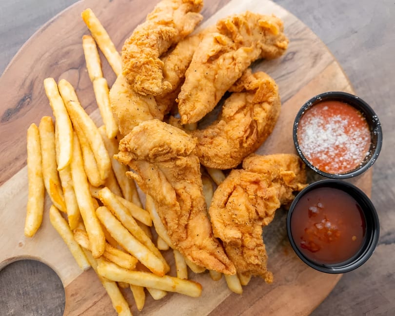 Crispy chicken tenders and golden french fries served with two dipping sauces on a wooden board.